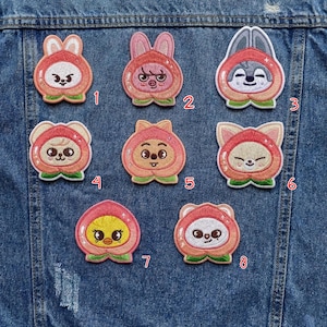 May include: A collection of eight embroidered patches featuring cartoon characters in peach-shaped costumes. Each patch has a unique character design with varying facial expressions and colors, set against a denim background.