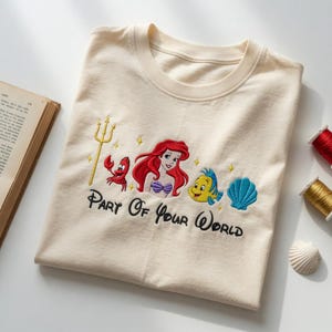 Puede incluir: Camiseta color crema con un dise&ntilde;o bordado de Ariel, Flounder, Sebasti&aacute;n y una concha. La frase "Part Of Your World" est&aacute; bordada en escritura negra debajo de los personajes. Un tridente dorado y estrellas completan el dise&ntilde;o.