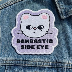 Pu&ograve; includere: Toppa ricamata con la faccia di un gatto bianco, orecchie rosa e occhi neri, su sfondo lilla chiaro. Il testo "BOMBASTIC SIDE EYE" &egrave; in nero. La toppa &egrave; cucita su una giacca di jeans.