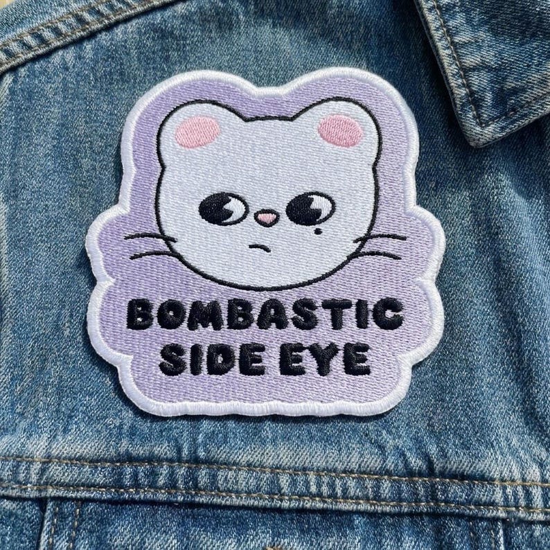 Pu&ograve; includere: Toppa ricamata con la faccia di un gatto bianco, orecchie rosa e occhi neri, su sfondo lilla chiaro. Il testo "BOMBASTIC SIDE EYE" &egrave; in nero. La toppa &egrave; cucita su una giacca di jeans.