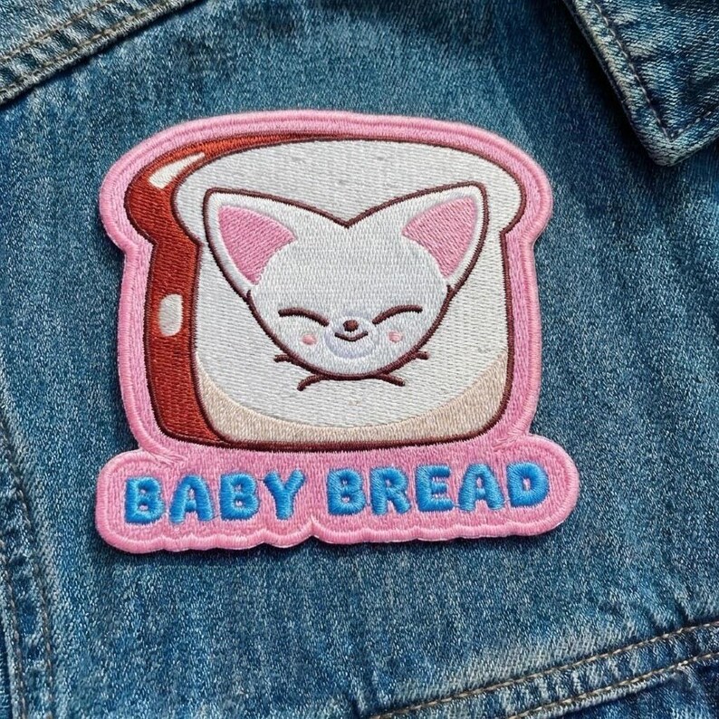 Pu&ograve; includere: Toppa ricamata con un volto di gatto dei cartoni animati su una fetta di pane. La toppa ha un bordo rosa e le parole "BABY BREAD" in lettere blu. La toppa &egrave; cucita su una giacca di jeans.