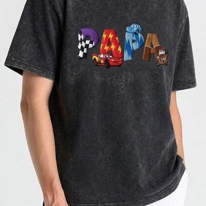 以下が含まれることがあります： 「PAPA」の文字がカラフルな漫画風の文字で描かれた、濃いグレーのTシャツ。各文字は、チェッカーフラッグ、稲妻、車など、異なるテーマでデザインされています。