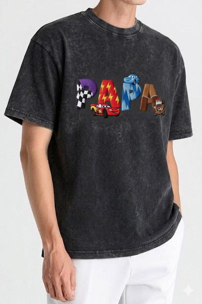以下が含まれることがあります： 「PAPA」の文字がカラフルな漫画風の文字で描かれた、濃いグレーのTシャツ。各文字は、チェッカーフラッグ、稲妻、車など、異なるテーマでデザインされています。