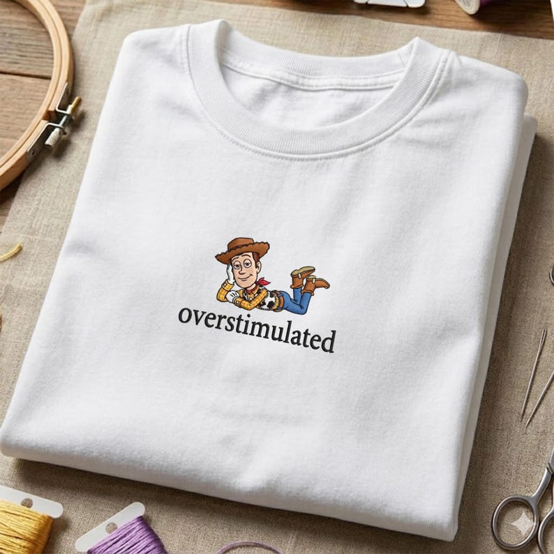 May include: White crew neck t-shirt with a cartoon cowboy character and the word "overstimulated" printed below. The cowboy wears a brown hat, yellow shirt, red bandana, blue jeans, and brown boots. The t-shirt is folded.