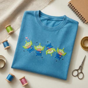 May include: A blue t-shirt featuring an embroidered design of four green three-eyed aliens in blue uniforms. The aliens are depicted in various poses, with two small pink hearts floating above the leftmost figure on the fabric.