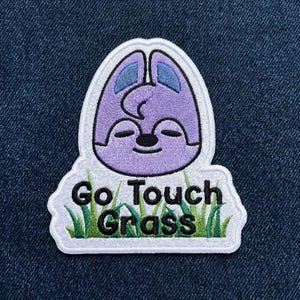 Pu&ograve; includere: Toppa ricamata con un personaggio viola dei cartoni animati con gli occhi chiusi e le orecchie blu. La toppa recita "Go Touch Grass" in testo nero e verde, con accenti di erba verde. Su sfondo denim blu.