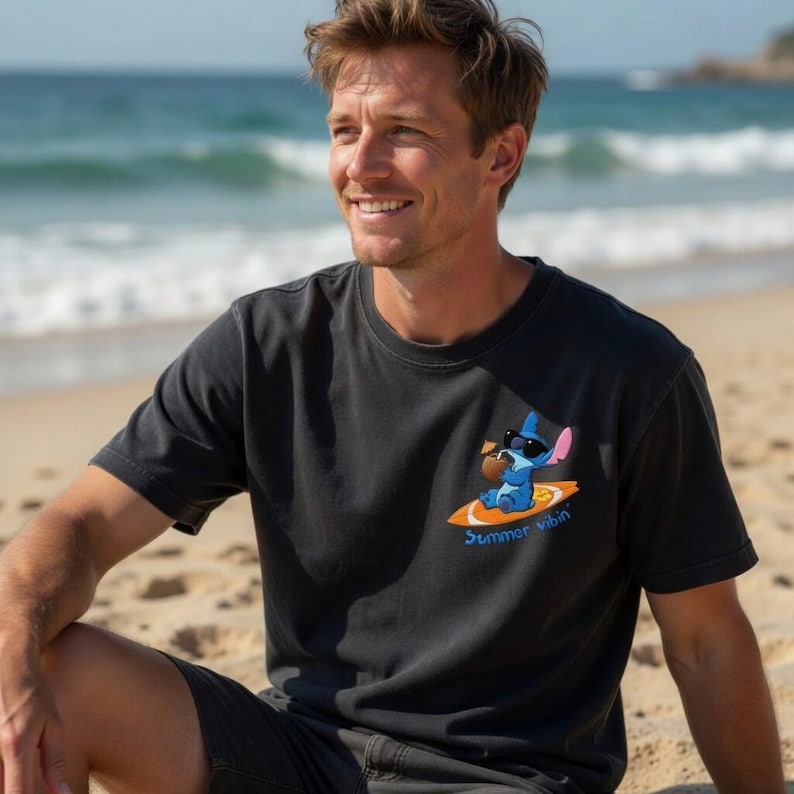 Op de afbeelding: Zwart t-shirt met een cartoonfiguur op een surfplank, met een zonnebril en een kokosdrankje. De tekst "Summer vibin'" staat eronder gedrukt. De persoon draagt het t-shirt op een zandstrand met de oceaan op de achtergrond.