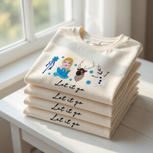 Puede incluir: Una pila de sudaderas de color crema con dise&ntilde;os bordados de personajes y la frase "Let it go". Los dise&ntilde;os incluyen una mujer rubia con un vestido azul, un reno y un mu&ntilde;eco de nieve. Las sudaderas est&aacute;n cuidadosamente dobladas sobre una superficie blanca.