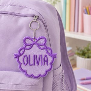May include: A lavender backpack featuring a circular embroidered keychain with a scalloped edge and a bow detail at the top. The keychain is attached with a silver metal ring and chain, and it displays the name OLIVIA in bold purple embroidery.