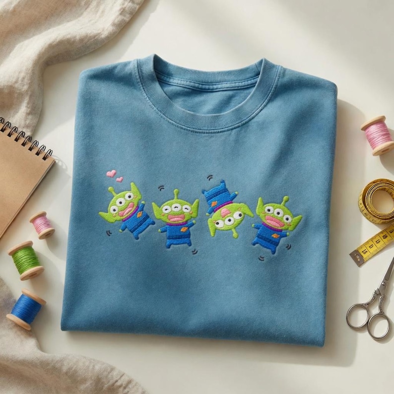 May include: A blue crew neck t-shirt featuring an embroidered design of four green three-eyed aliens in blue uniforms. The aliens are depicted in various playful poses, with one upside down and two small pink hearts floating above one alien.