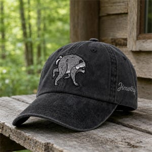 Peut inclure: Une casquette de baseball gris anthracite avec une broderie représentant un raton laveur penché sur le panneau avant. Le côté de la casquette affiche le nom Jimothy brodé en écriture cursive. La casquette possède une visière courbée et des œillets.