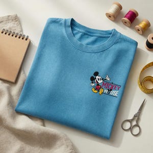 K&ouml;nnte beinhalten: Hellblaues T-Shirt mit gesticktem Mickey Mouse-Design und dem Text "Mickey Mouse". Das Bild zeigt N&auml;hzubeh&ouml;r: Garnrollen, Schere und ein Ma&szlig;band, alles auf einer wei&szlig;en Oberfl&auml;che.