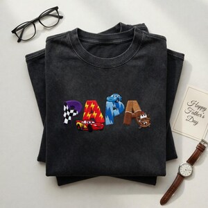 以下が含まれることがあります： カラフルな文字で「PAPA」と刺繍された濃いグレーのTシャツ。それぞれ異なる車のデザインが特徴です。「Happy Father's Day」と書かれたカードも表示されています。