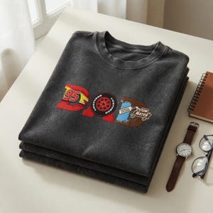 May include: A stack of three charcoal gray t-shirts with a graphic design featuring the word "DAD" in a Cars movie theme. The design includes the number "95", a tire, and the words "Tow Mater". A watch, glasses, and a notebook are also visible.