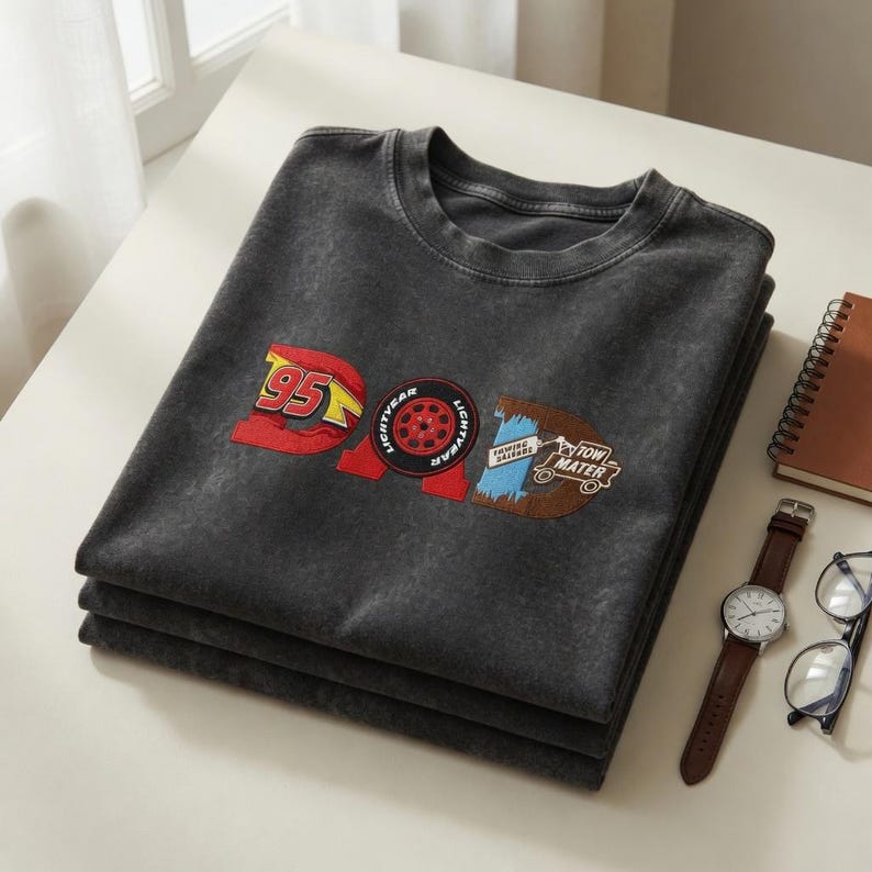May include: A stack of three charcoal gray t-shirts with a graphic design featuring the word "DAD" in a Cars movie theme. The design includes the number "95", a tire, and the words "Tow Mater". A watch, glasses, and a notebook are also visible.