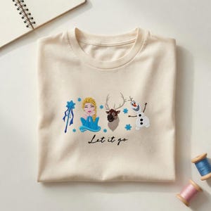 Puede incluir: Camiseta color crema con bordados de personajes de la pel&iacute;cula Frozen. El dise&ntilde;o incluye a Elsa, Sven, Olaf y la frase "Let it go". Acentos de copos de nieve azules completan el dise&ntilde;o.