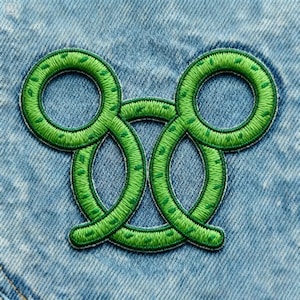Könnte beinhalten: Ein grüner, gestickter Aufnäher mit einem sich wiederholenden Acht-Muster. Der Aufnäher ist mit leuchtend grünem Faden genäht und hat ein strukturiertes Aussehen. Der Aufnäher ist auf einem hellblauen Denim-Stoff befestigt.