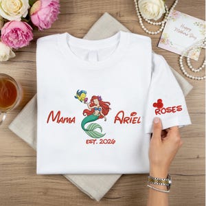 May include: White t-shirt featuring a colorful embroidered design of Ariel from The Little Mermaid, with the words "Mama Ariel" and "EST. 2026" in red. The sleeve has a red embroidered Mickey Mouse head and the word "Roses".