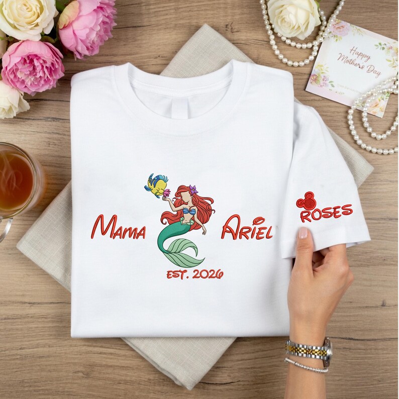 May include: White t-shirt featuring a colorful embroidered design of Ariel from The Little Mermaid, with the words "Mama Ariel" and "EST. 2026" in red. The sleeve has a red embroidered Mickey Mouse head and the word "Roses".
