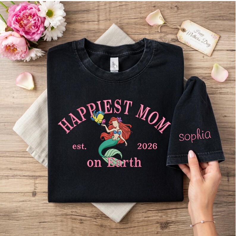 May include: Black t-shirt with the text "Happiest Mom on Earth" in pink, featuring a mermaid graphic. The shirt also has the text "est. 2026". A matching black sleeve with the name "Sophia" is also visible.