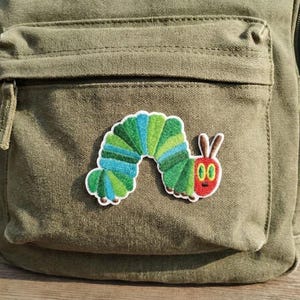 May include: An olive green canvas backpack with a colourful embroidered patch of a cartoon caterpillar. The caterpillar patch features green, blue, and red colours. The backpack has a front pocket and zip.