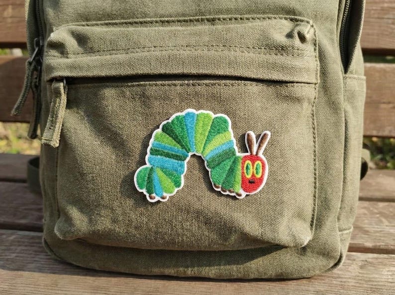 May include: An olive green canvas backpack with a colourful embroidered patch of a cartoon caterpillar. The caterpillar patch features green, blue, and red colours. The backpack has a front pocket and zip.