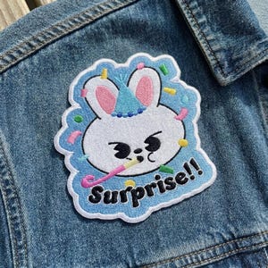 Pu&ograve; includere: Toppa ricamata con un coniglietto bianco con cappello da festa, orecchie rosa ed espressione sorpresa. La toppa ha un bordo blu con la scritta "Surprise!!" in nero e dettagli di coriandoli. Su una giacca di jeans.
