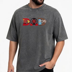 May include: Dark gray t-shirt with the word "DAD" in a graphic design. The "D" is a red "95" with a yellow lightning bolt, the "A" is a tire, and the second "D" is a brown design with the words "TOW MATER".