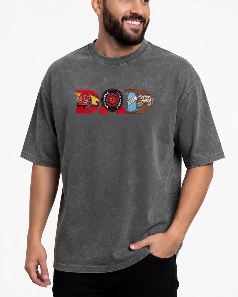 May include: Dark gray t-shirt with the word "DAD" in a graphic design. The "D" is a red "95" with a yellow lightning bolt, the "A" is a tire, and the second "D" is a brown design with the words "TOW MATER".