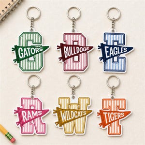 May include: Six embroidered keychains featuring large block letters with vertical stripes, each overlaid with a pennant flag displaying team names: GATORS, BULLDOGS, EAGLES, RAMS, WILDCATS, and TIGERS. Each keychain includes a silver metal ring and chain.