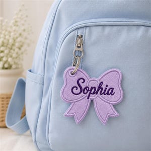 May include: A light blue backpack featuring a purple embroidered bow-shaped keychain attached to the front zipper. The keychain displays the name Sophia in dark purple cursive script, centered on the fabric bow with a silver metal ring.