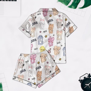 May include: White satin pajama set featuring a repeating pattern of cartoon animals in pastel colors. The set includes a short-sleeved button-up shirt and matching shorts. The text "SKZ" is printed throughout the design.