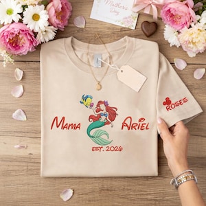 May include: Beige t-shirt featuring a colorful illustration of Ariel from The Little Mermaid with the text "Mama Ariel EST. 2026". The sleeve has the word "ROSES" embroidered in red. A gold necklace and a blank tag are also visible.