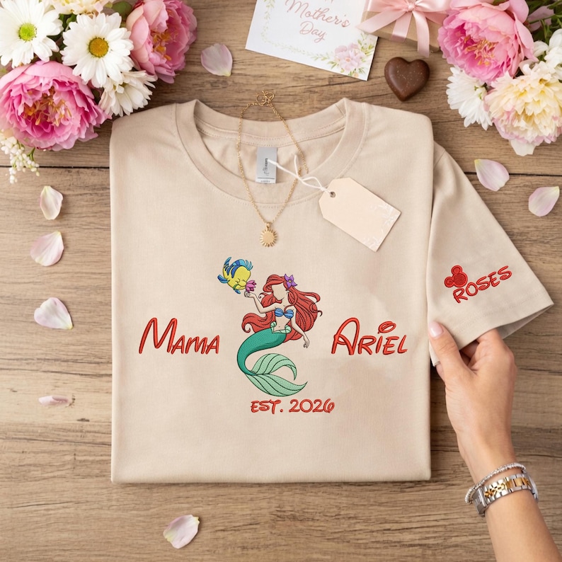 May include: Beige t-shirt featuring a colorful illustration of Ariel from The Little Mermaid with the text "Mama Ariel EST. 2026". The sleeve has the word "ROSES" embroidered in red. A gold necklace and a blank tag are also visible.
