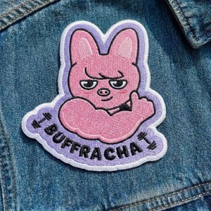Pu&ograve; includere: Toppa ricamata raffigurante un coniglio rosa con un braccio muscoloso che fa il pollice in su. La toppa ha un bordo viola e la parola "BUFFRACHA" &egrave; ricamata intorno al coniglio. La toppa &egrave; su una giacca di jeans blu.