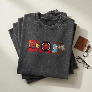 May include: A stack of charcoal gray t-shirts with the word "DAD" embroidered in red, blue, and brown. The "D" features the number "95" and the "A" is a tire. The "D" has the words "Tow Mater". A brown notebook, watch, and glasses are next to the shirts.