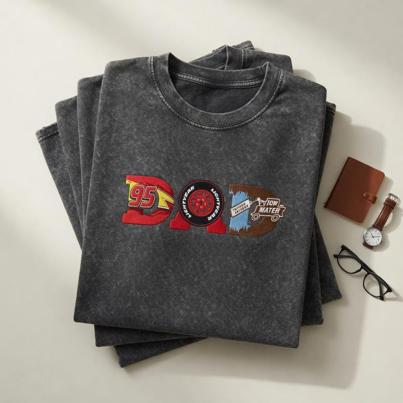May include: A stack of charcoal gray t-shirts with the word "DAD" embroidered in red, blue, and brown. The "D" features the number "95" and the "A" is a tire. The "D" has the words "Tow Mater". A brown notebook, watch, and glasses are next to the shirts.