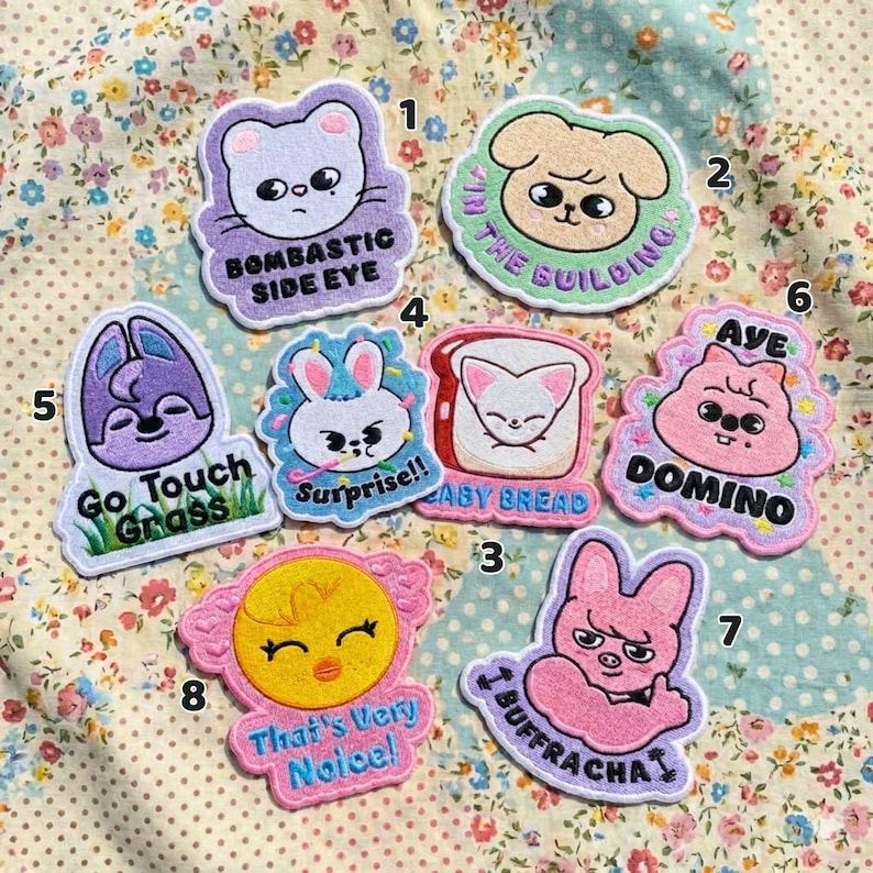Pu&ograve; includere: Una collezione di otto toppe ricamate colorate con disegni di animali e cibo dei cartoni animati. Le toppe presentano testi come "Bombastic Side Eye", "Baby Bread" e "That's Very Noice!" su uno sfondo a motivi floreali.