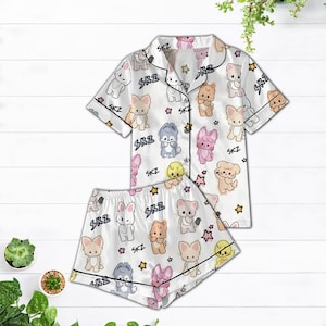May include: White satin pajama set featuring a repeating pattern of cartoon animals and stars. The short-sleeved top and shorts have black trim and the letters "SKZ" are printed throughout the design. The set is displayed on a white wooden surface.