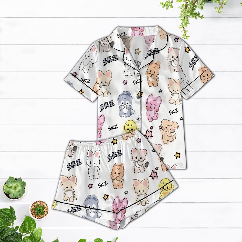 May include: White satin pajama set featuring a repeating pattern of cartoon animals and stars. The short-sleeved top and shorts have black trim and the letters "SKZ" are printed throughout the design. The set is displayed on a white wooden surface.