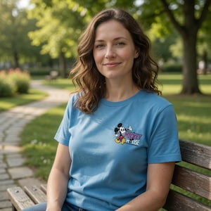 K&ouml;nnte beinhalten: Hellblaues T-Shirt mit Mickey Mouse-Grafik. Die Grafik zeigt Mickey Mouse und die Worte "Mickey Mouse". Das T-Shirt ist kurz&auml;rmlig und aus weichem Material. Die Person sitzt auf einer Holzbank.