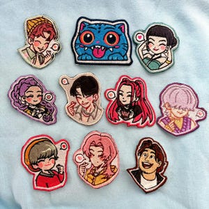 Puede incluir: Una colección de parches bordados con personajes de estilo caricatura. Los parches muestran una variedad de diseños, incluyendo personas y un gato azul con una amplia sonrisa. Cada parche tiene un diseño detallado y colorido con un borde cosido.