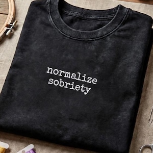 Può includere: T-shirt nero con le parole bianche "normalize sobriety" centrate sul davanti. La maglietta ha uno scollo tondo e un aspetto leggermente sbiadito. L'immagine è ripresa dall'alto.