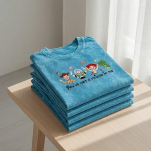 Puede incluir: Una pila de cuatro camisetas turquesas con cuello redondo. Cada camiseta presenta un dise&ntilde;o bordado de personajes de dibujos animados populares, con el texto "You've got a friend in me" en letra cursiva.