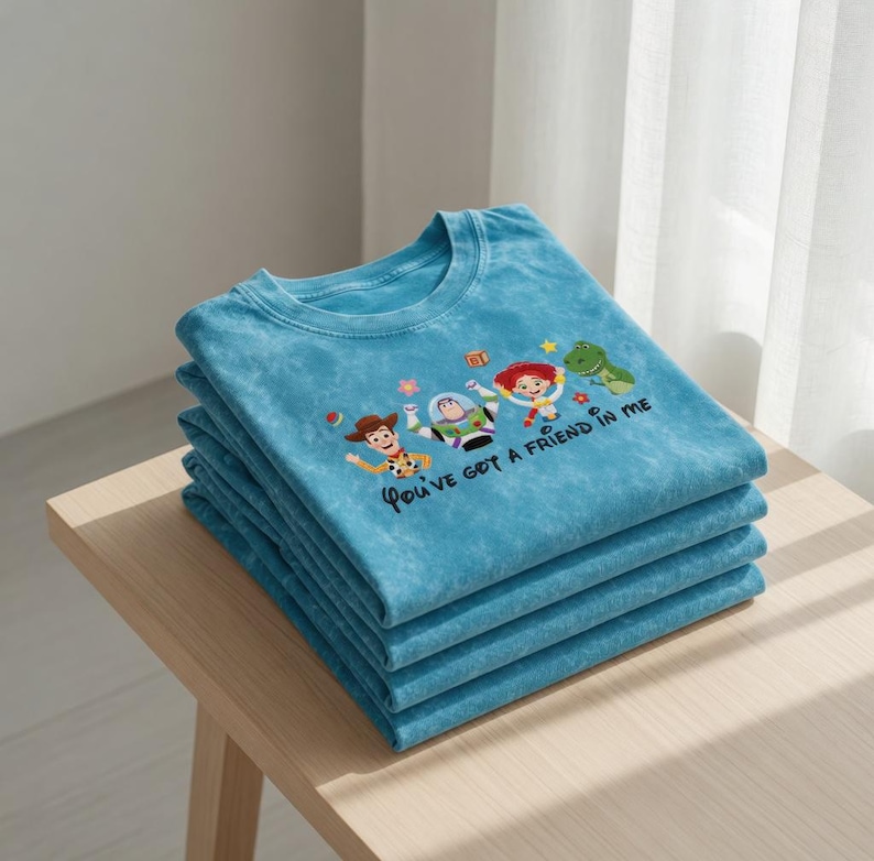 Puede incluir: Una pila de cuatro camisetas turquesas con cuello redondo. Cada camiseta presenta un dise&ntilde;o bordado de personajes de dibujos animados populares, con el texto "You've got a friend in me" en letra cursiva.