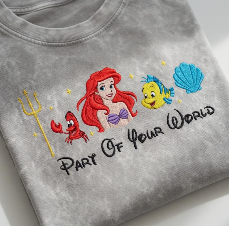 Puede incluir: Camiseta gris con un dise&ntilde;o bordado de Ariel, Flounder, Sebasti&aacute;n y una concha azul. La frase "Part Of Your World" est&aacute; bordada debajo de los personajes. Un tridente dorado y estrellas amarillas completan el dise&ntilde;o.