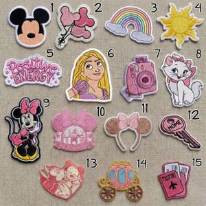 Op de afbeelding: Een verzameling geborduurde patches met diverse ontwerpen. Inclusief Mickey Mouse, ballonnen, een regenboog, een zon, de tekst "POSITIVE ENERGY", Rapunzel, een roze camera, Marie de kat, Minnie Mouse, een Disneyland-embleem, Minnie Mouse oren, een sleutel met "HOME", Mickey en Minnie, een koets en paspoorten.