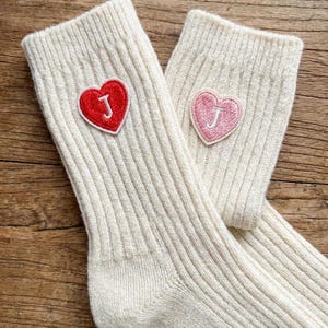 Puede incluir: Un par de calcetines de punto acanalado de color crema. Cada calcetín tiene un parche en forma de corazón con la letra "J" bordada. Un corazón es rojo, el otro es rosa.