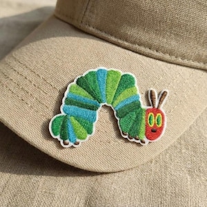 May include: A beige baseball cap with a vibrant embroidered caterpillar patch. The caterpillar design includes green, blue, and red colours, with a white border. The patch is sewn onto the front of the cap.
