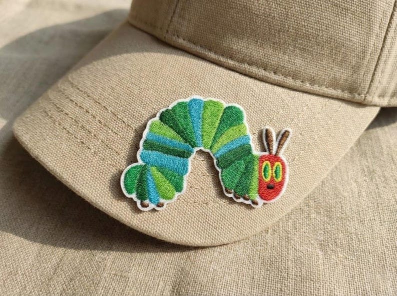 May include: A beige baseball cap with a vibrant embroidered caterpillar patch. The caterpillar design includes green, blue, and red colours, with a white border. The patch is sewn onto the front of the cap.
